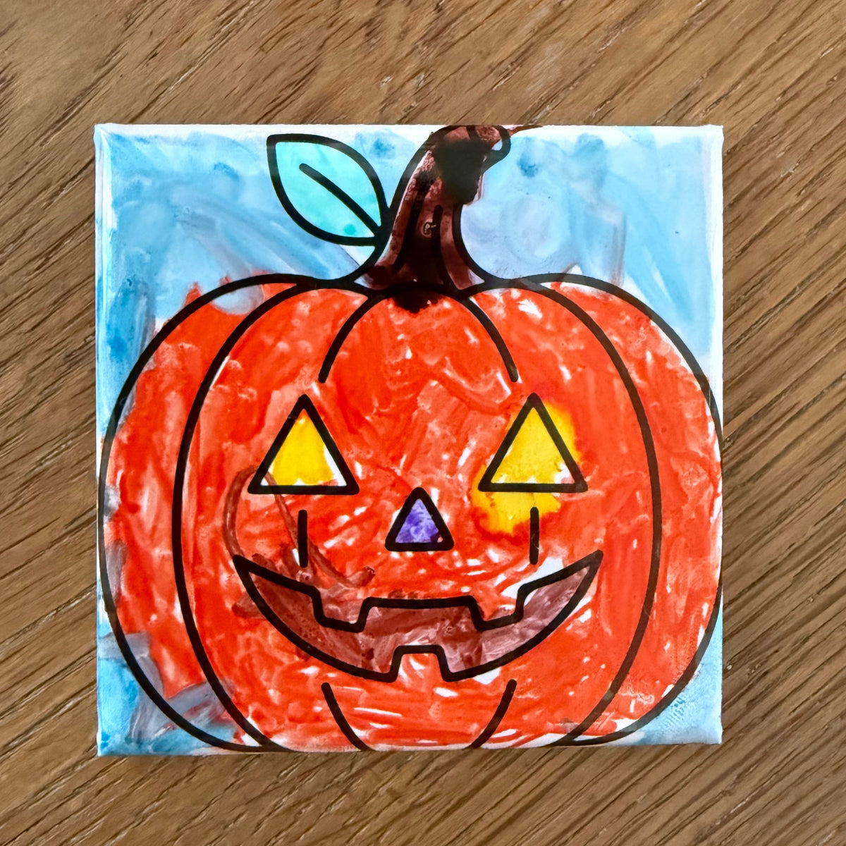 A square ceramic tile featuring a hand-painted jack-o'-lantern pumpkin design, placed on a brown wooden surface.